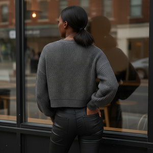 Person wearing a gray textured sweater and black pants standing by a window with a cityscape view.