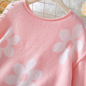 Pink sweater with white floral patterns on a hanger against a blurred floral background