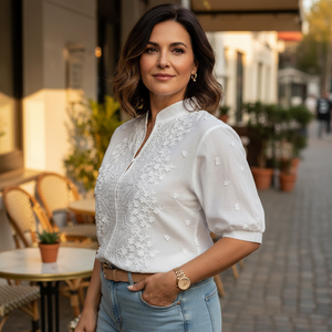 Woman in a white blouse with floral patterns standing on a street with outdoor seating.