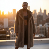 Woman in a fur coat standing on a rooftop with a cityscape in the background during sunset.