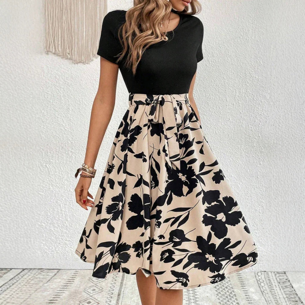 Woman wearing a black top and floral skirt against a white wall.