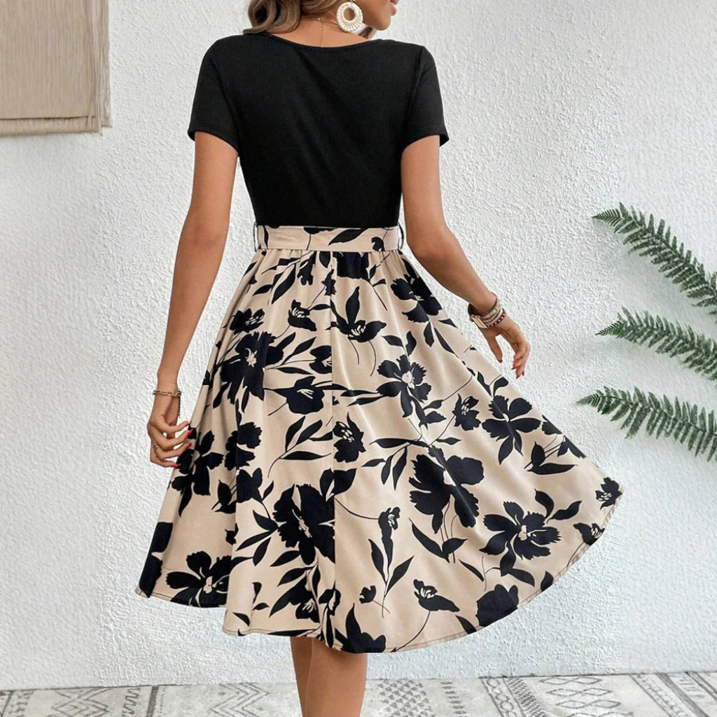 Woman wearing a black top and floral skirt against a white wall.