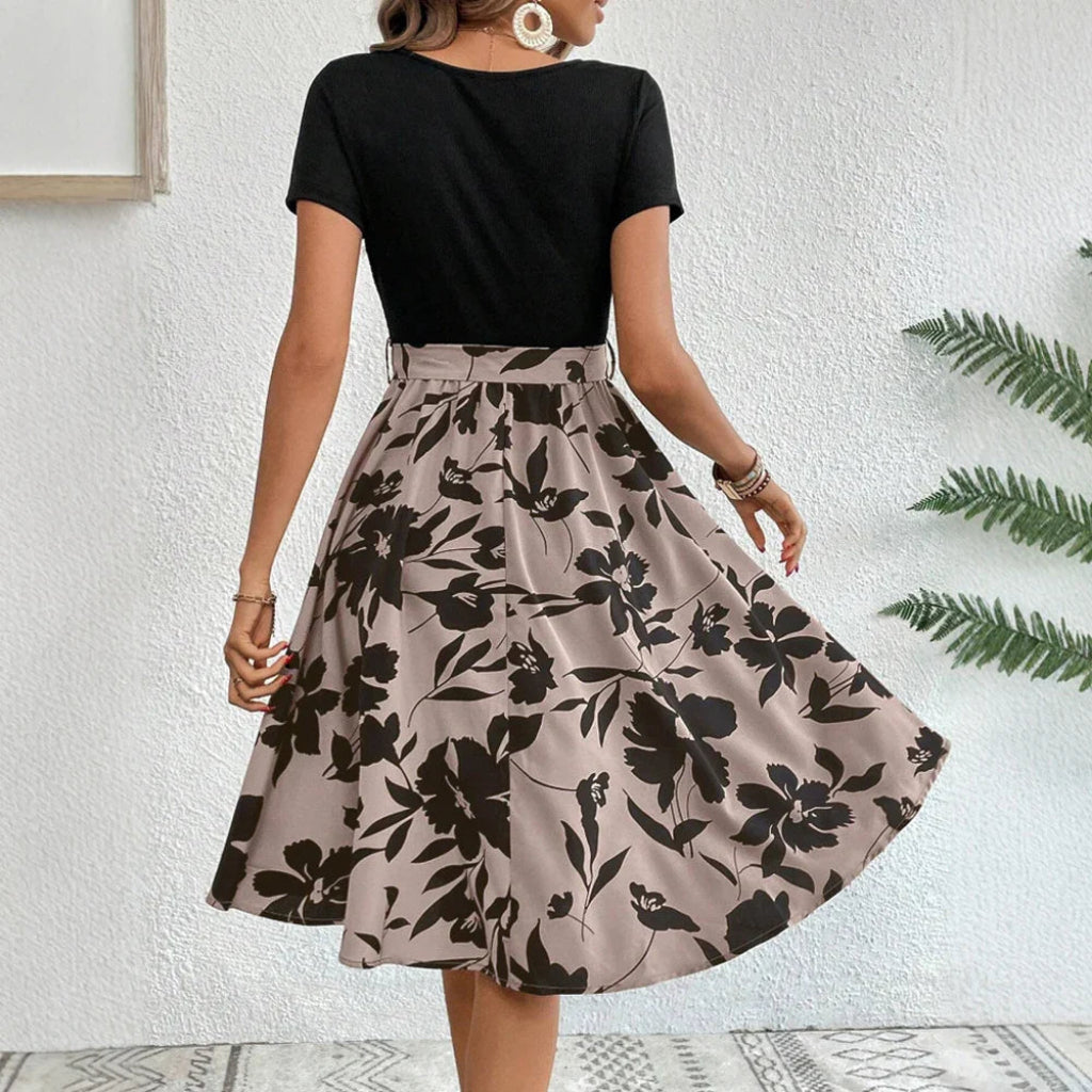 Woman wearing a black top and floral skirt against a white wall.