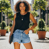 Woman wearing a black sleeveless top and denim shorts standing outdoors with plants and buildings in the background.