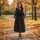 Woman in a long black puffer coat standing on a path with autumn leaves and trees in the background