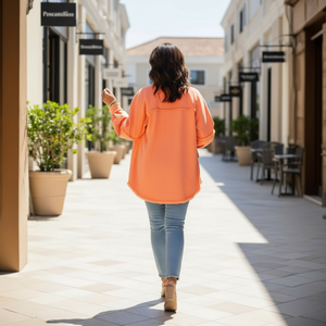 Woman in an orange jacket walking on a sunlit street with shops and plants.