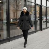 Woman in a black coat with fur collar walking on a city sidewalk.
