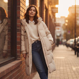 Woman in a long beige coat standing on a city street with warm lighting.