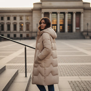 Woman in a long beige coat standing in front of a large building with columns.