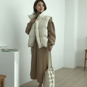 Woman wearing a beige puffer vest over a brown outfit, holding a tote bag indoors.