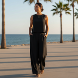 Woman in a black sleeveless top and wide-leg pants walking on a beach with palm trees in the background.