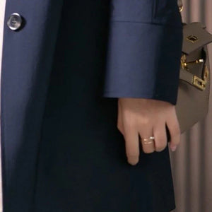 Person wearing a navy coat with a hand holding a beige handbag, wearing a gold ring.