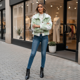 Woman wearing a light green jacket, white shirt, blue jeans, and black boots standing on a sidewalk in front of a store.