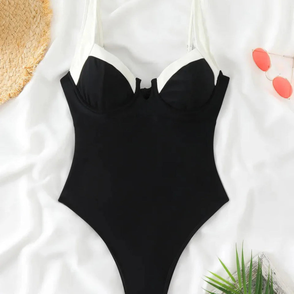 Black one-piece swimsuit on a white surface with a straw hat and green leaves.