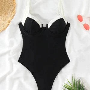 Black one-piece swimsuit on a white surface with a straw hat and green leaves.