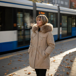 Woman wearing a beige parka with fur hood standing at a tram stop in autumn.