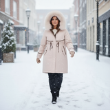 Woman walking in the snow wearing a long beige coat with a fur-lined hood.