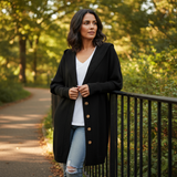 Woman wearing a black coat and light blue jeans standing on a path with greenery.
