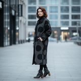 Woman wearing a long black coat with large polka dots on a city street.