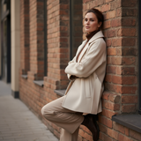 Woman in a beige coat leaning against a brick wall.